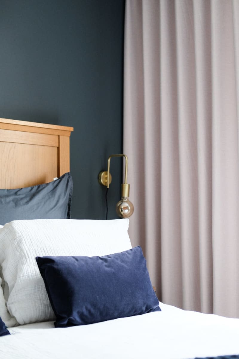 slate gray walls with light wood headboard, gold sconce fixture and white linens with dark blue accent pillows, cream curtains