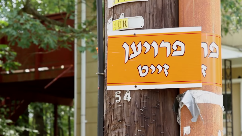 Sign with Hebrew text attached to a utility pole, with foliage and a building in the background