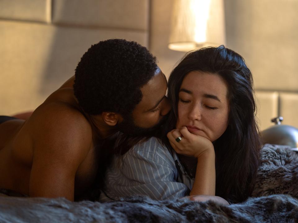 donald glover and maya erskine in mr. and mrs. smith, laying together in bed. glover is shirtless, pressing his nose into erskine's neck as she looks back at him