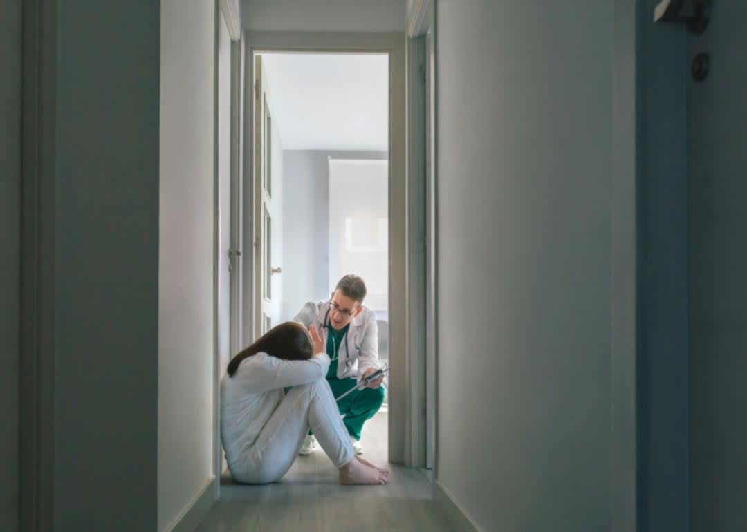 A psychiatric tech kneeling down to help a patient sitting on the floor.