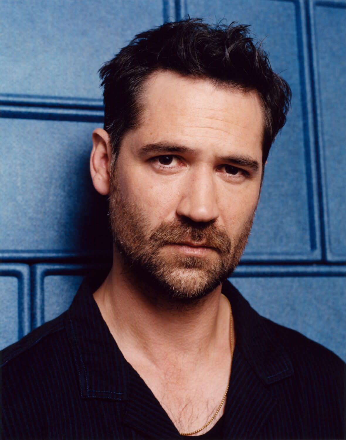 A man wearing a black shirt looks at the camera while leaning against a blue wall.