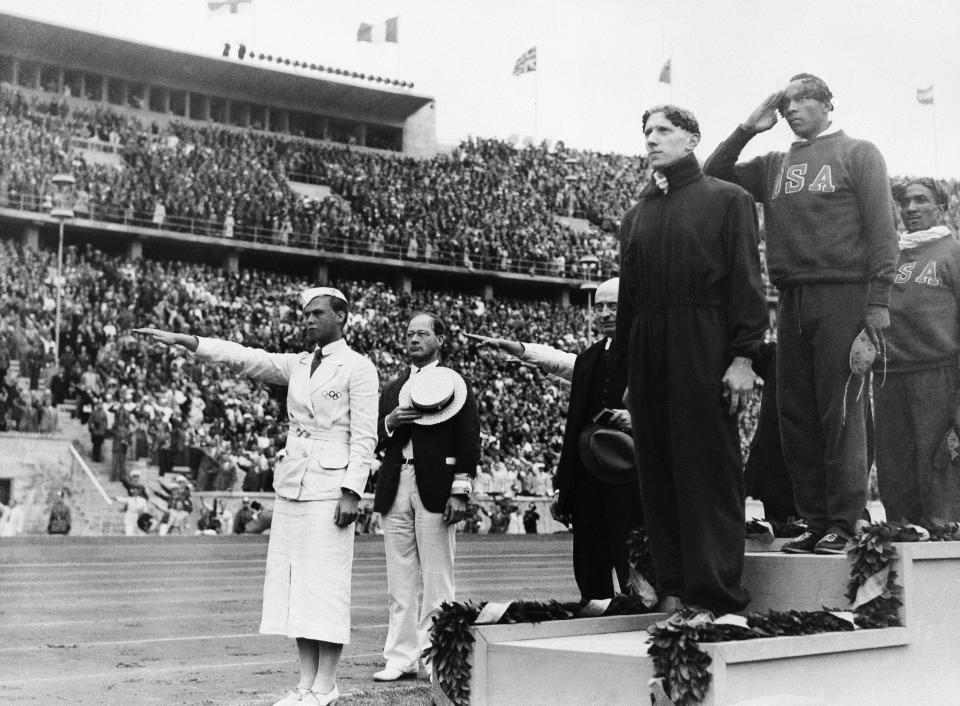 FILE - In this Aug. 3, 1936, file photo, gold medalist Jessie Owens, second right, salutes during the playing of the national anthem during the medal ceremony of the 100-meter final in Berlin. silver medalist Tinus Osendarp, third from right, of, Holland, and bronze medalist Ralph Metcalf, right, listen, along with a matron who holds a Nazi salute. The 1936 Games in Berlin, awarded about two years before Adolf Hitler became dictator, went ahead under Nazism. American track great Jesse Owens went on to win four gold medals, but he was only supposed to compete in three events, the 100 meters, 200 meters and long jump. (AP Photo/File)