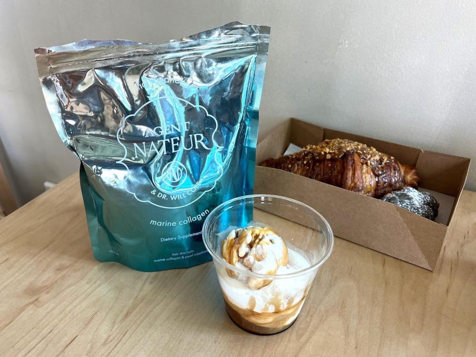 A blue metallic bag of marine collagen, a cup with coffee and a scoop of ice cream inside, and a box of breakfast pastries on a table.