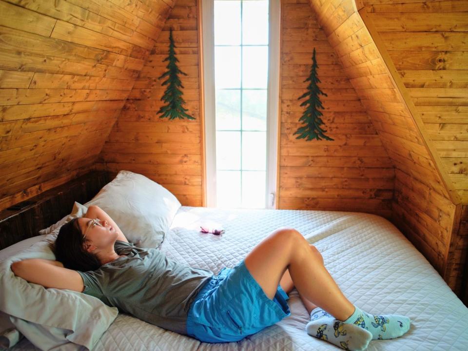 the author laying in the home's white bed looking out the window. The walls are wooden and there are two decals of pine trees on either sidee of the window.