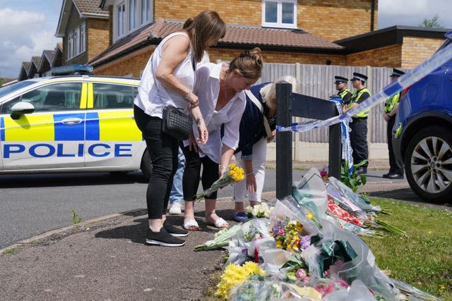 Three women place flowers at a street sign. There are lots of other tributes, a police car is in the background with two police officers. There is police crime scene tape visible at the top of the image