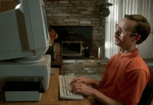 A man with glasses and a mustache sits at a desk, typing on a large, old-fashioned computer in a room with a brick fireplace