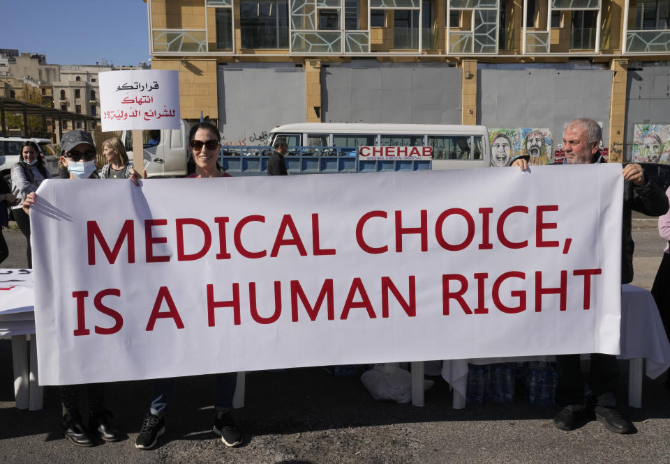 Protesters hold a banner during a rally to protest measures imposed against people who are not vaccinated, in Beirut, Lebanon, Saturday, Jan. 8, 2022. Vaccinations are not compulsory in Lebanon but in recent days authorities have become more strict in dealing with people who are not inoculated or don't carry a negative PCR test. Arabic placard on the left,reads: "Your decisions is a violation of international law." (AP Photo/Hussein Malla)