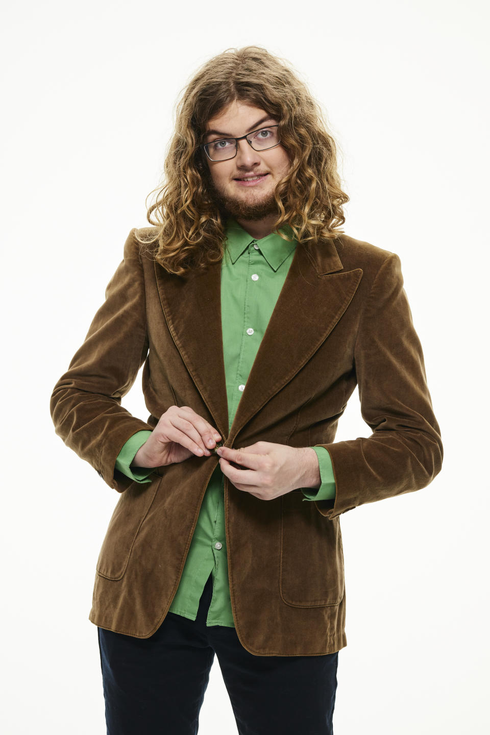 A man with long curly brown hair wears a brown velvet jacket over a bright green shirt and dark trousers. 