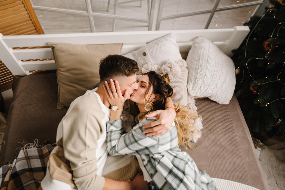 Man kisses and hugs woman lying on bed near Christmas tree in decorated bedroom interior. Couple enjoying winter time together on holidays. Happy New Year and Merry Christmas. Xmas at home. Top view.