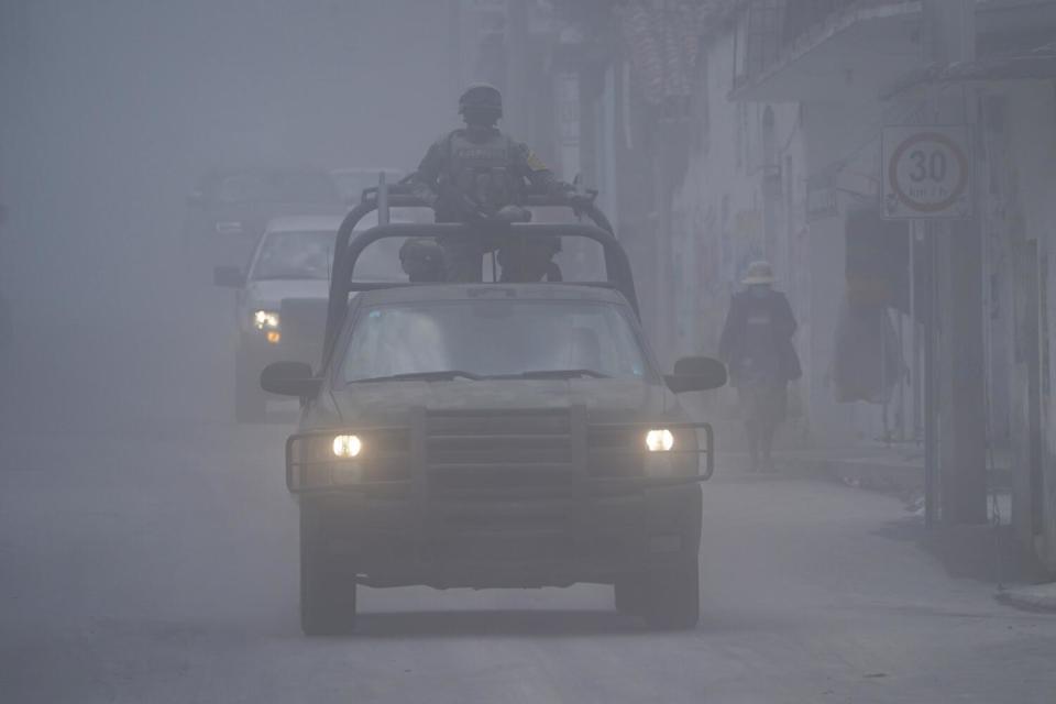 A person stands in the back of one of two vehicles driving with headlights on along an ash-covered street