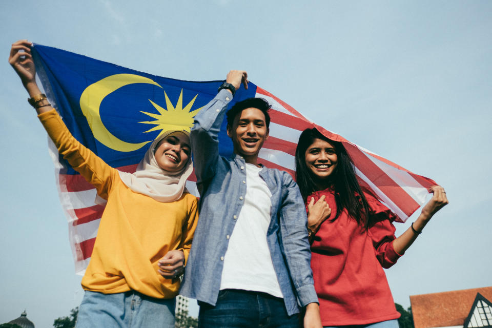 A malay lady, chinese man, and an indian lady holding the Malaysia flag over their heads.