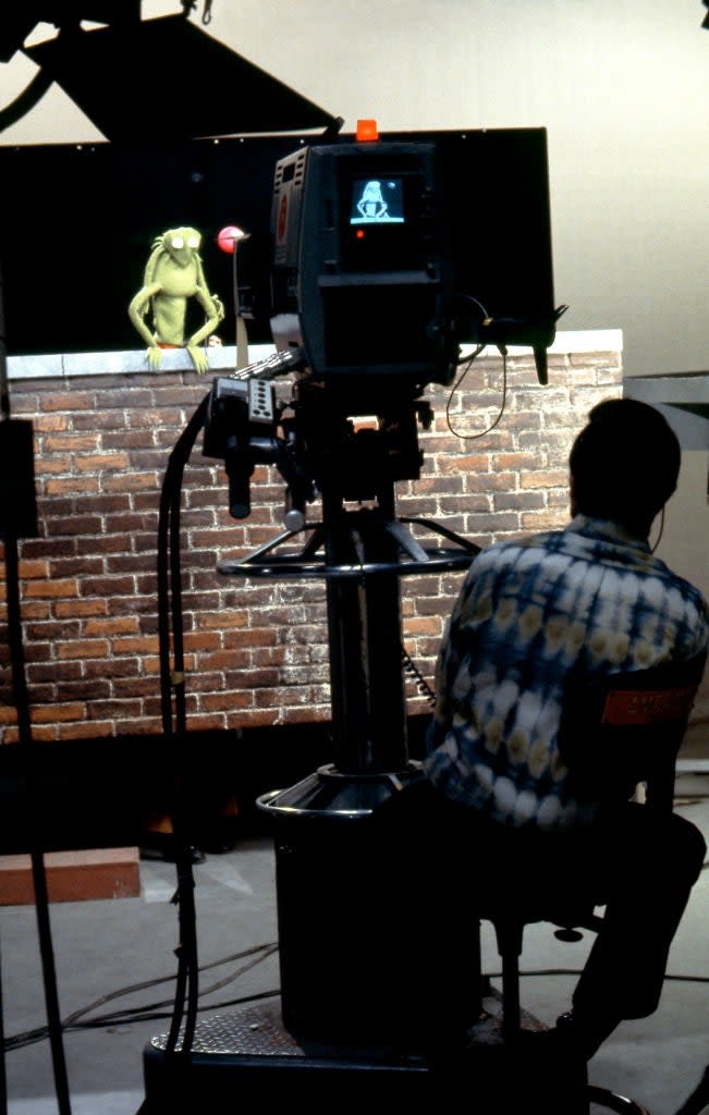 A puppeteer operates Kermit the Frog behind a set, while a cameraman films the scene