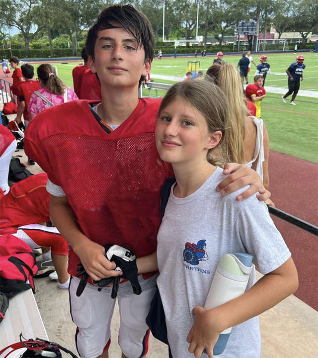 Benjamin Brady in a red shirt at a football game/practice stands tall next to his sister Viivan in a white t-shirt