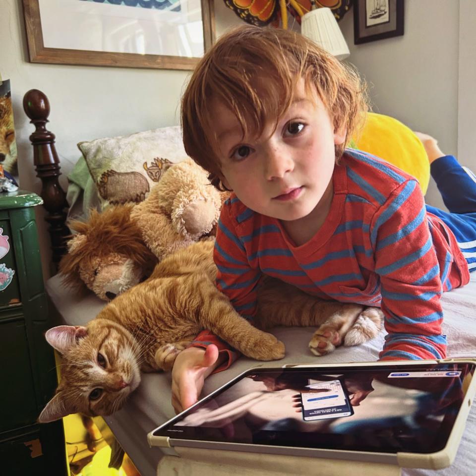 Melissa Petro's son snuggling with a cat and looking at an iPad screen on a bed.