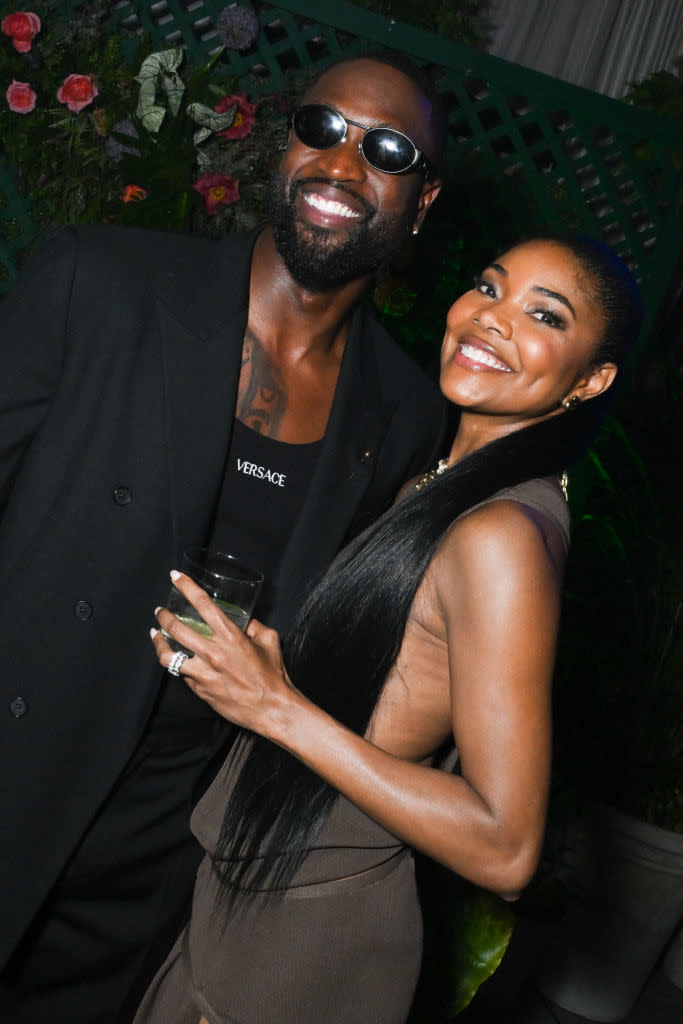 Two people smiling, one wearing a suit with Versace logo, the other with a long dress and ponytail