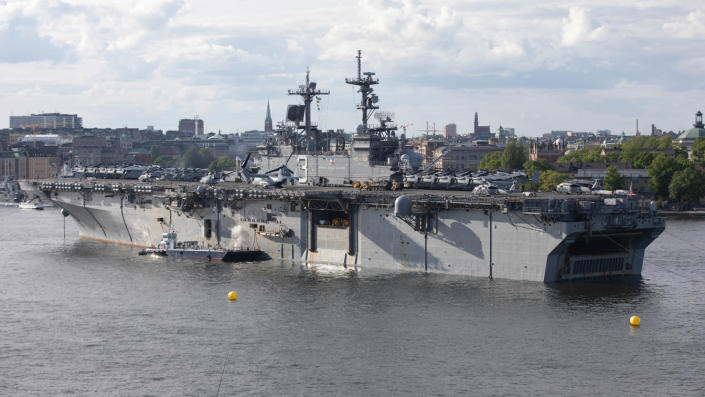 Large warship beneath cloudy skies against a background of a city skyline.