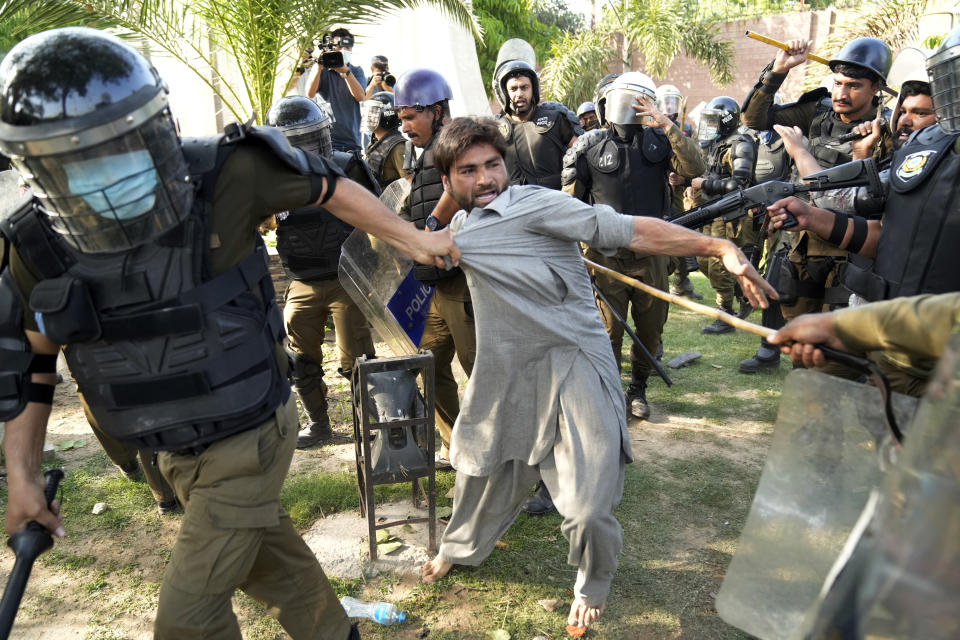 A helmeted officer drags a man wearing a cotton outfit by pulling on his shirt, as another officer points a long bamboo switch at the man.