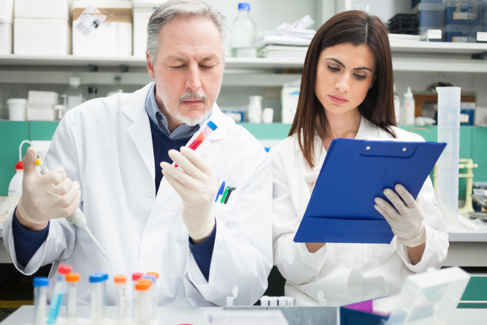 A man and woman in white lab coats stand side by side in a lab, examining a clipboard and the contents of several test tubes.