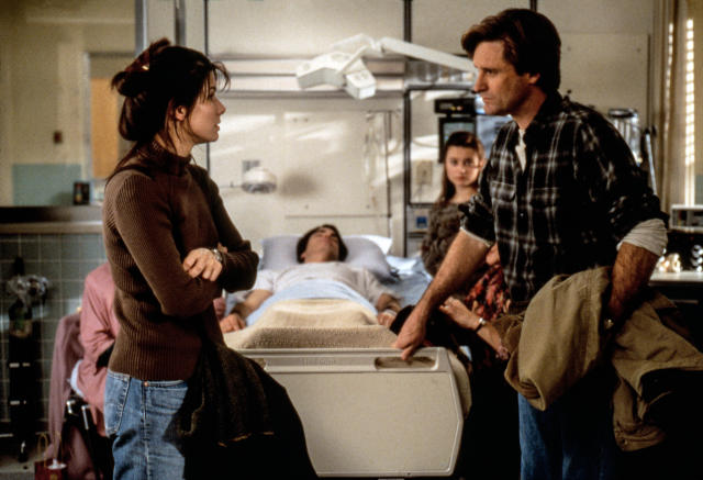 Sandra Bullock and Bill Pullman talk standing in front of a hospital bed on which a patient is lying