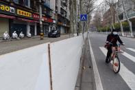 Woman wearing a face mask rides a shared bicycle past workers in protective suits who are resting in front of closed shops sealed off from the road, in Wuhan