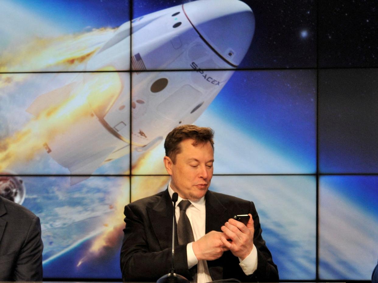 Elon Musk looks at his phone with a display of a space capsule entering orbit behind him.