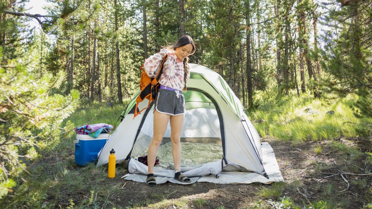  Young woman at campsite putting on backpack. 
