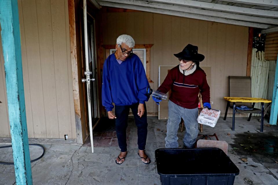 Two men stand on a concrete floor; one in a brimmed hat holds a plastic bottle in one hand and toilet paper in the other.