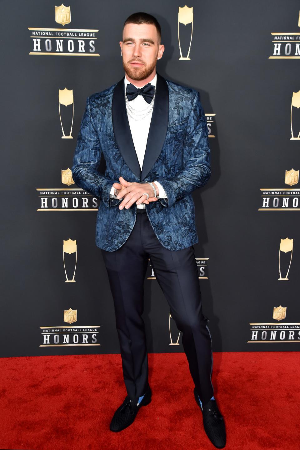 NFL player Travis Kelce wearing a patterned blue suit jacket, a bow-tie, and navy slacks on the red carpet at the NFL Honors.