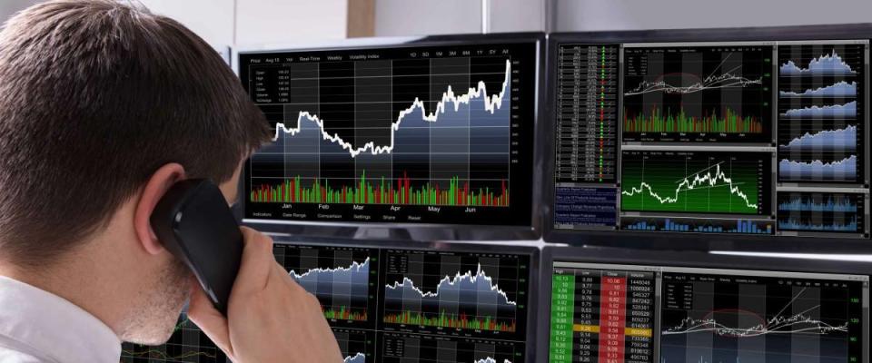 Stock Broker Talking On Telephone In Front Of Multiple Computer Screen Showing Graphs