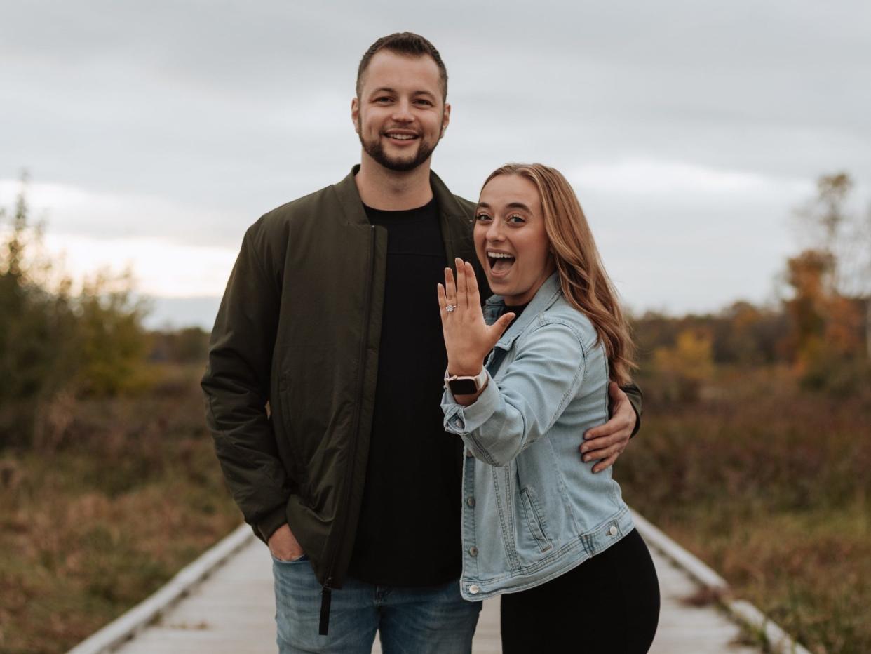 A man and woman on a boardwalk with the woman showing off an engagement ring.