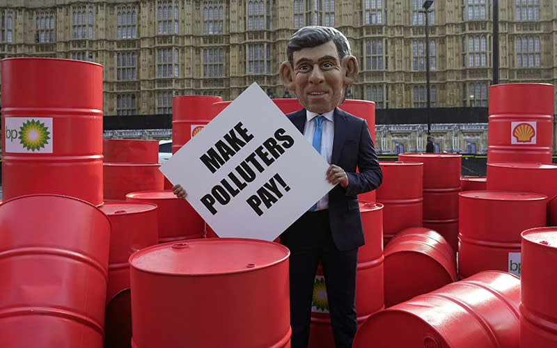 An Oxfam protester wearing a big head mask to resemble Rishi Sunak protests outside the Parliament. The man is wearing a suit and holding a sign that reads "MAKE POLLUTERS PAY!" He is standing in the center of the image, surrounded by red oil drums that have the logos of oil companies such as Shell and BP.