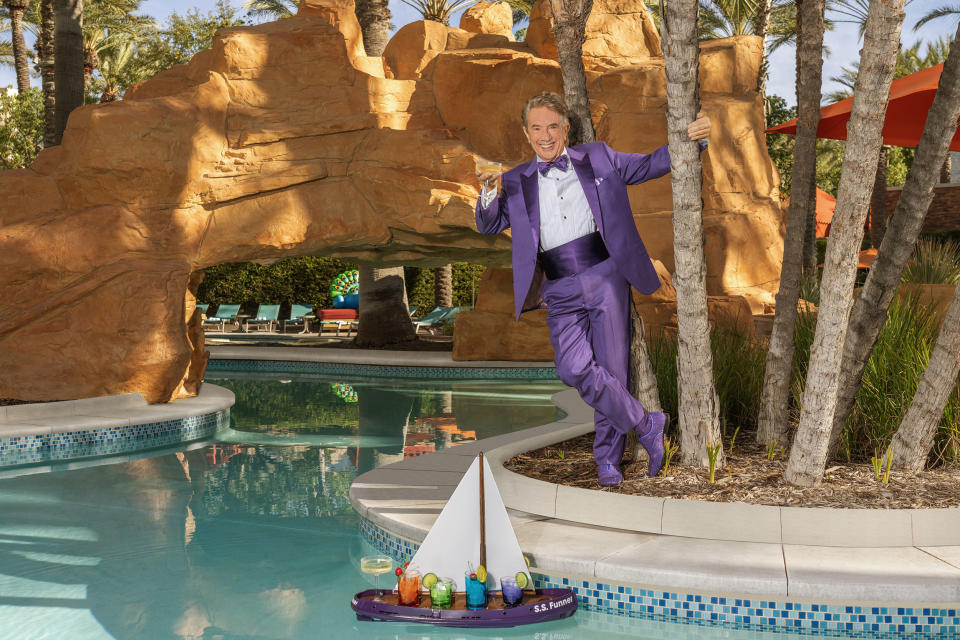 Martin Short dressed in a purple tuxedo holds on to a palm tress and poses by a swimming pool with a drink in his hand.