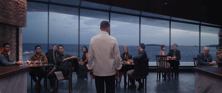 Ralph Fiennes, his back to the camera, looks out as his clean, modern restaurant. The wall behind is actually a floor to ceiling window, showing a rugged, bare landscape outside.