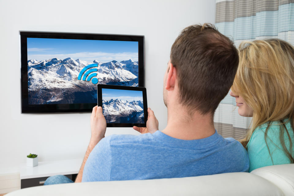 A couple sitting on a couch, watching a TV, and holding a tablet.