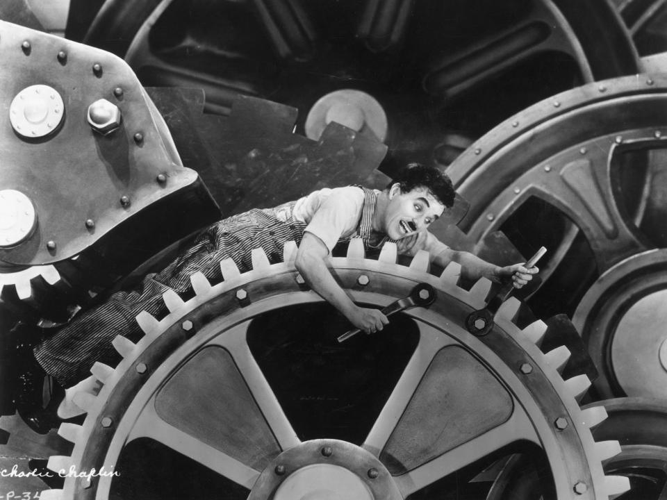 Charles Chaplin wearing overalls and holding a wrench, sits on an enormous set of gears.