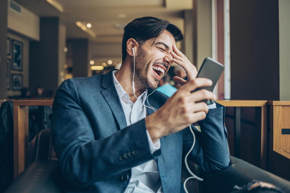 Young man with smartphone and headphones sitting in cafe and laughing