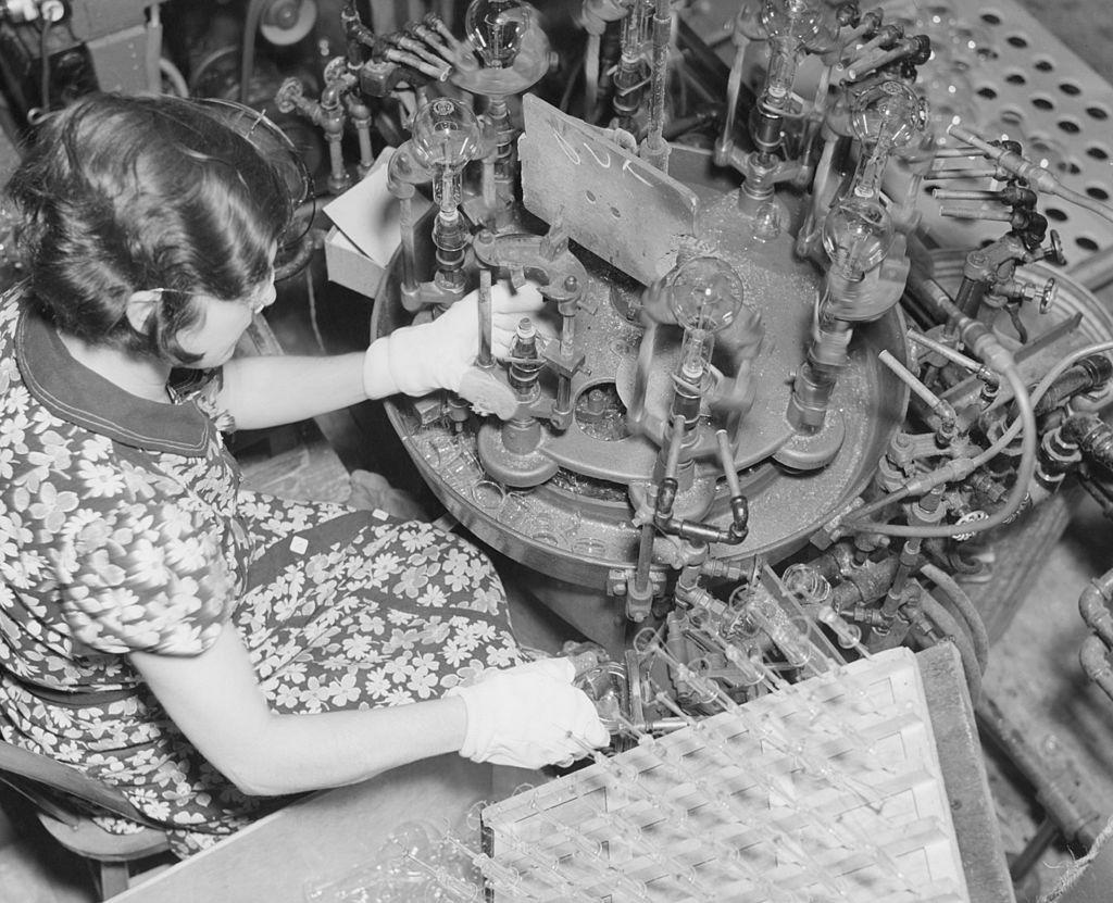 A female worker, wearing white gloves, 'sealing in' a light bulb on a production line, at a light bulb factory, 