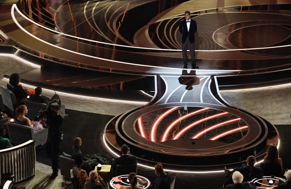 Presenter Chris Rock speaks onstage as Will Smith and Jada Pinkett, bottom left, listen from their seats at the Oscars, Sunday, March 27, 2022, at the Dolby Theatre in Los Angeles. (AP Photo/Chris Pizzello)