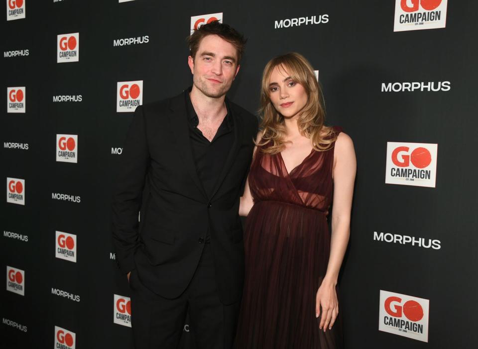 robert pattinson and suki waterhouse pose on the carpet, he's wearing a black suit and has an arm around her waist, she's wearing a see through burgundy dress