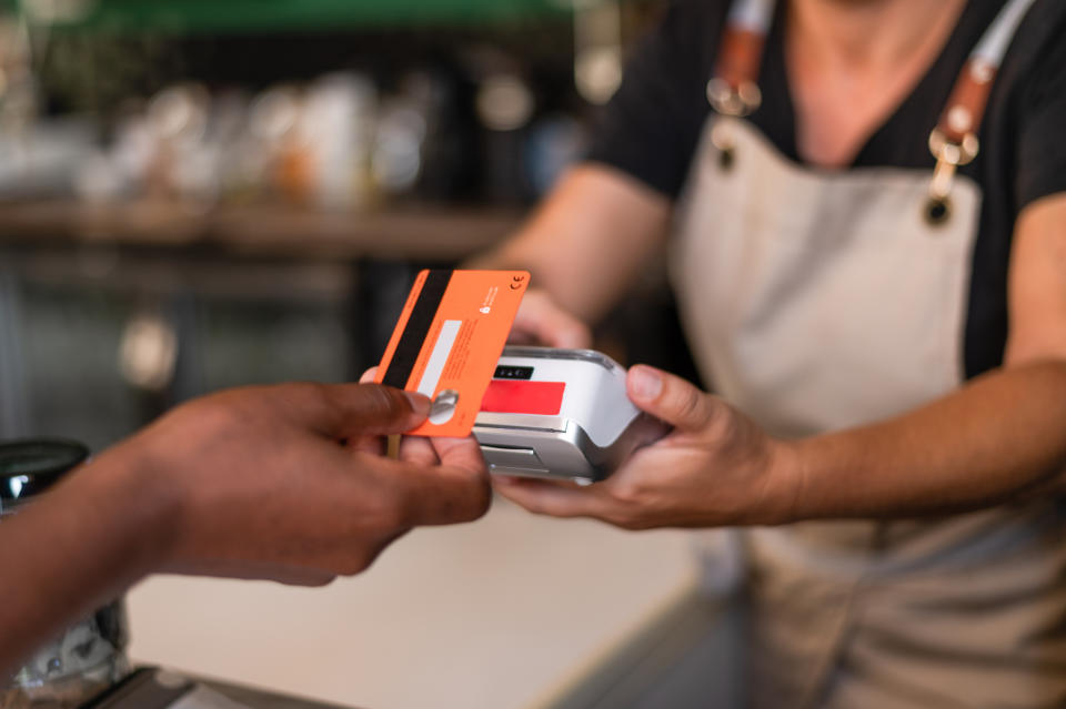 A person wearing an apron holds a payment terminal while another hand holds a credit card, preparing to complete a transaction at a counter