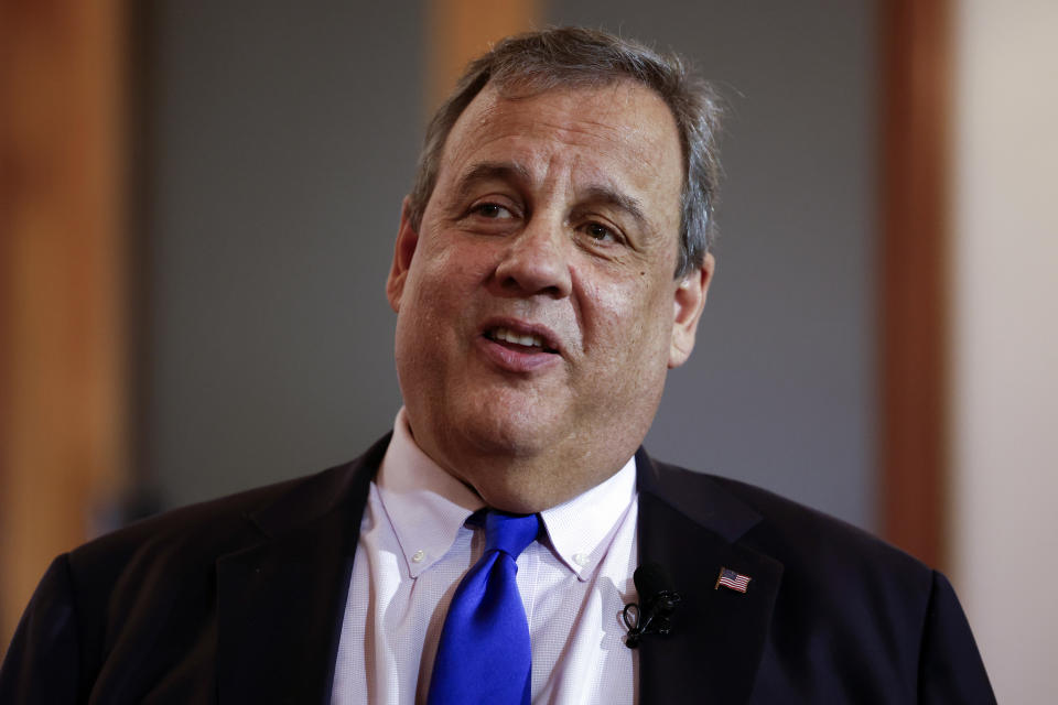 Individual speaking at an event, wearing a suit and tie with an American flag lapel pin