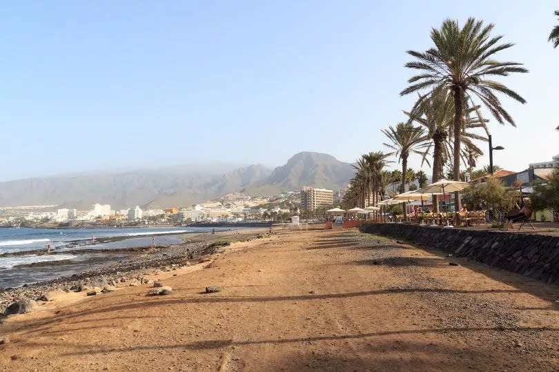 A golden sand beach with a promenade on the right with parasols and tables.