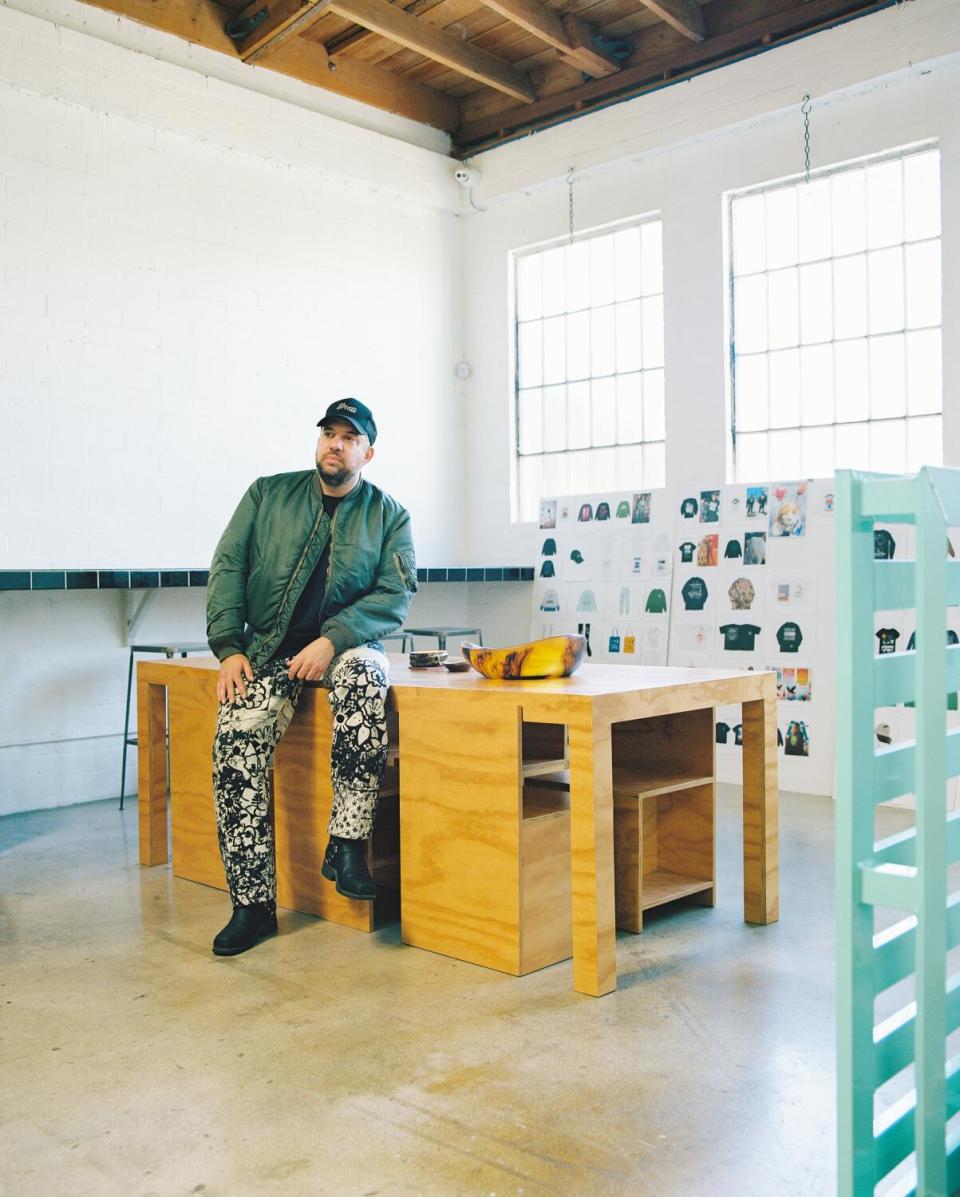 A man wearing patterned pants and a cap sits on a desk.