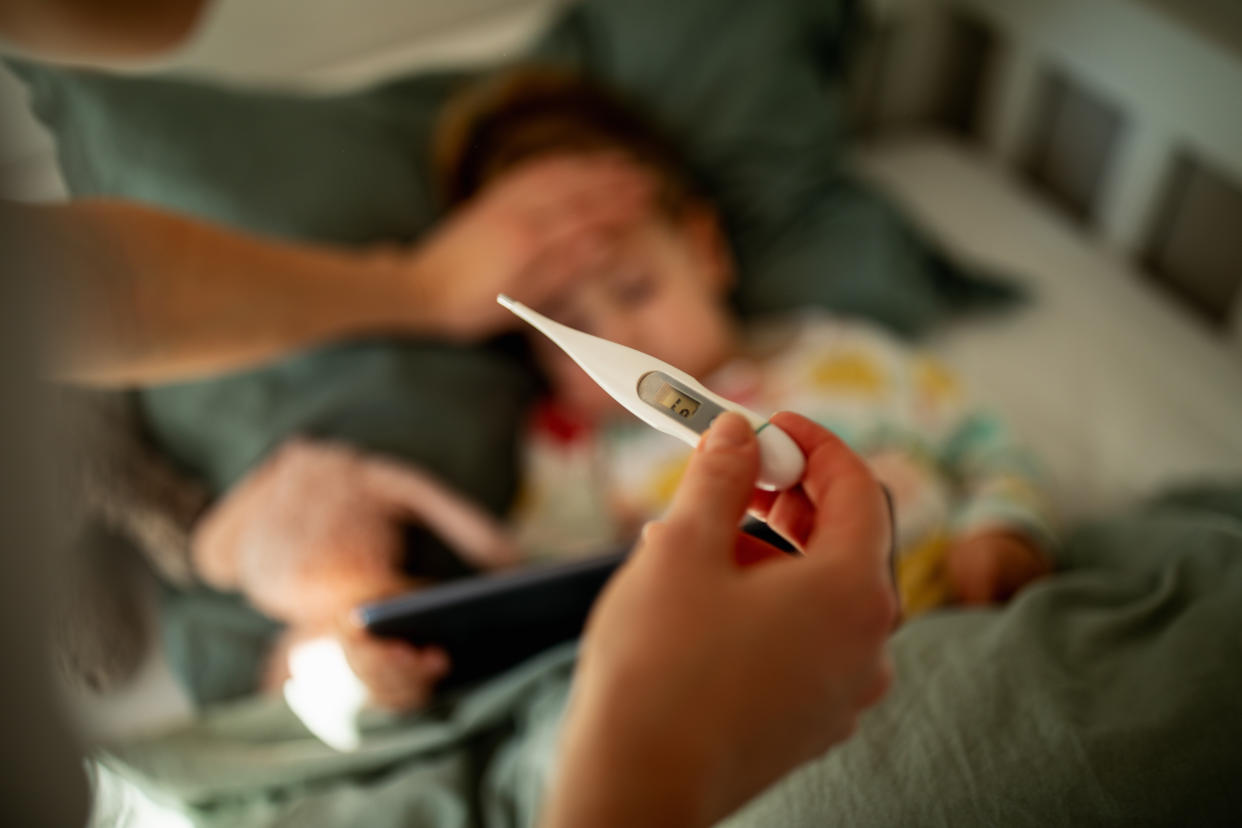 Close up of mother taking care of her sick child in cozy bright and warm kids bedroom with pastel green background in sunny morning light. Little girl has a cold and her mother is measuring her temperature with digital thermometer .