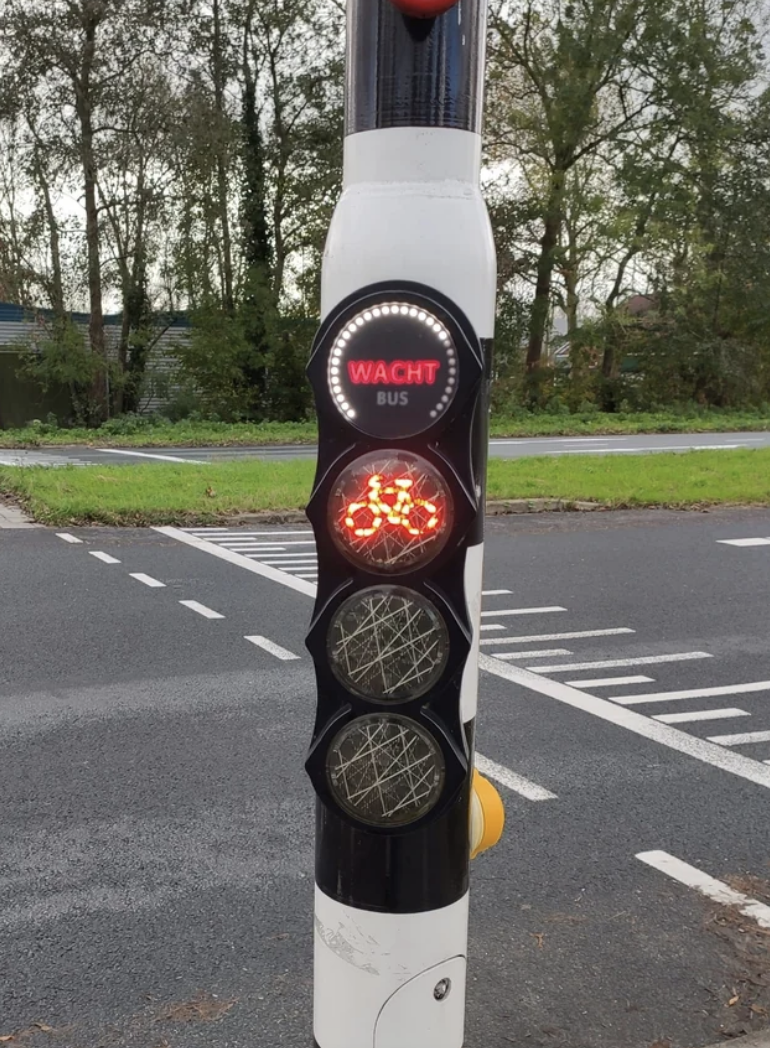 Traffic light for buses displaying a red light with "WACHT" sign illuminated, indicating wait, next to a road