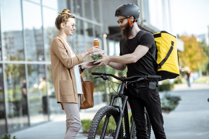 A food delivery worker on a bicycle handing an order to a customer.