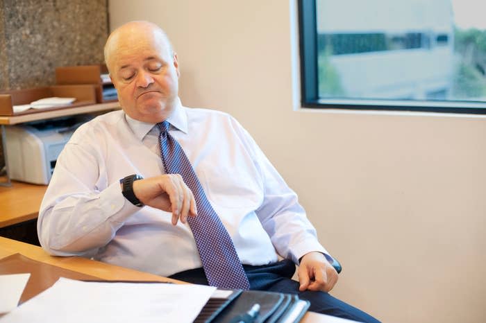 A middle-aged man in business attire checks his wristwatch, sitting at a desk in an office with a window in the background