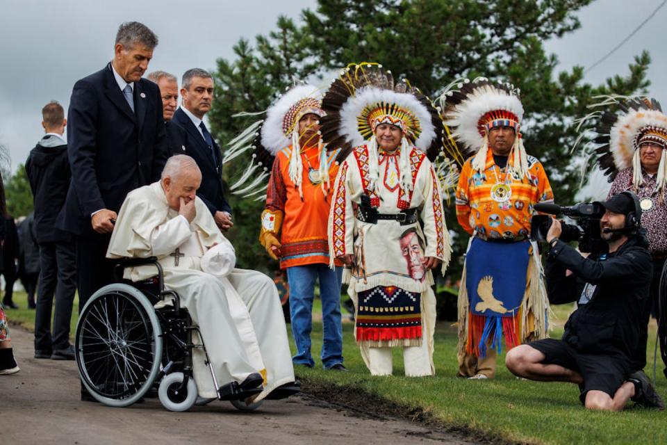 The pope sits in a wheelchair, his hand to his face, while three men in headdresses stand nearby.