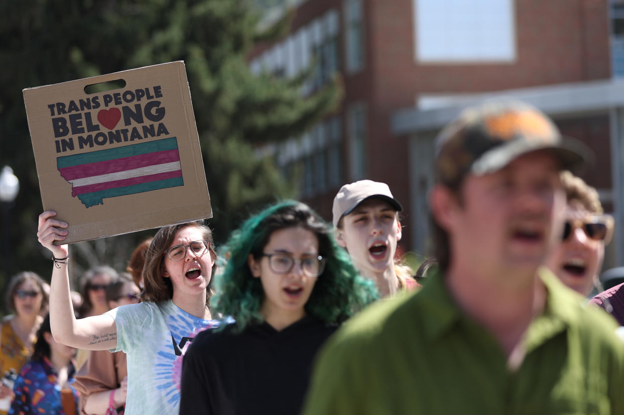 Transgender rights activists on the march, chanting. One young woman has green hair, and one holds a poster saying: Trans People Belong in Montana, with a map of the state bisected by green, red and white stripes.. 