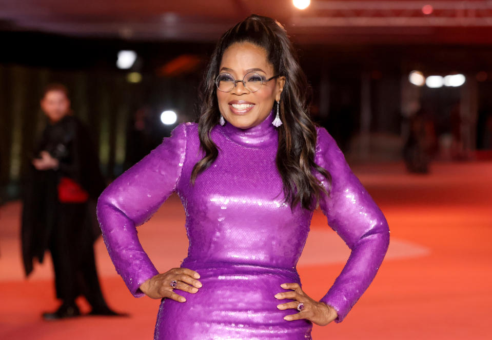 Oprah Winfrey smiles in a long-sleeved, sequined purple dress with glasses on the red carpet